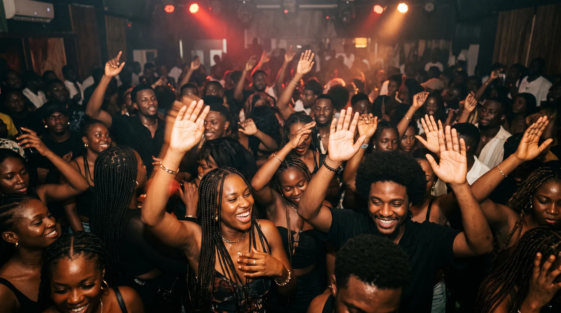 A packed dance floor of guests with arms raised under red and amber lights